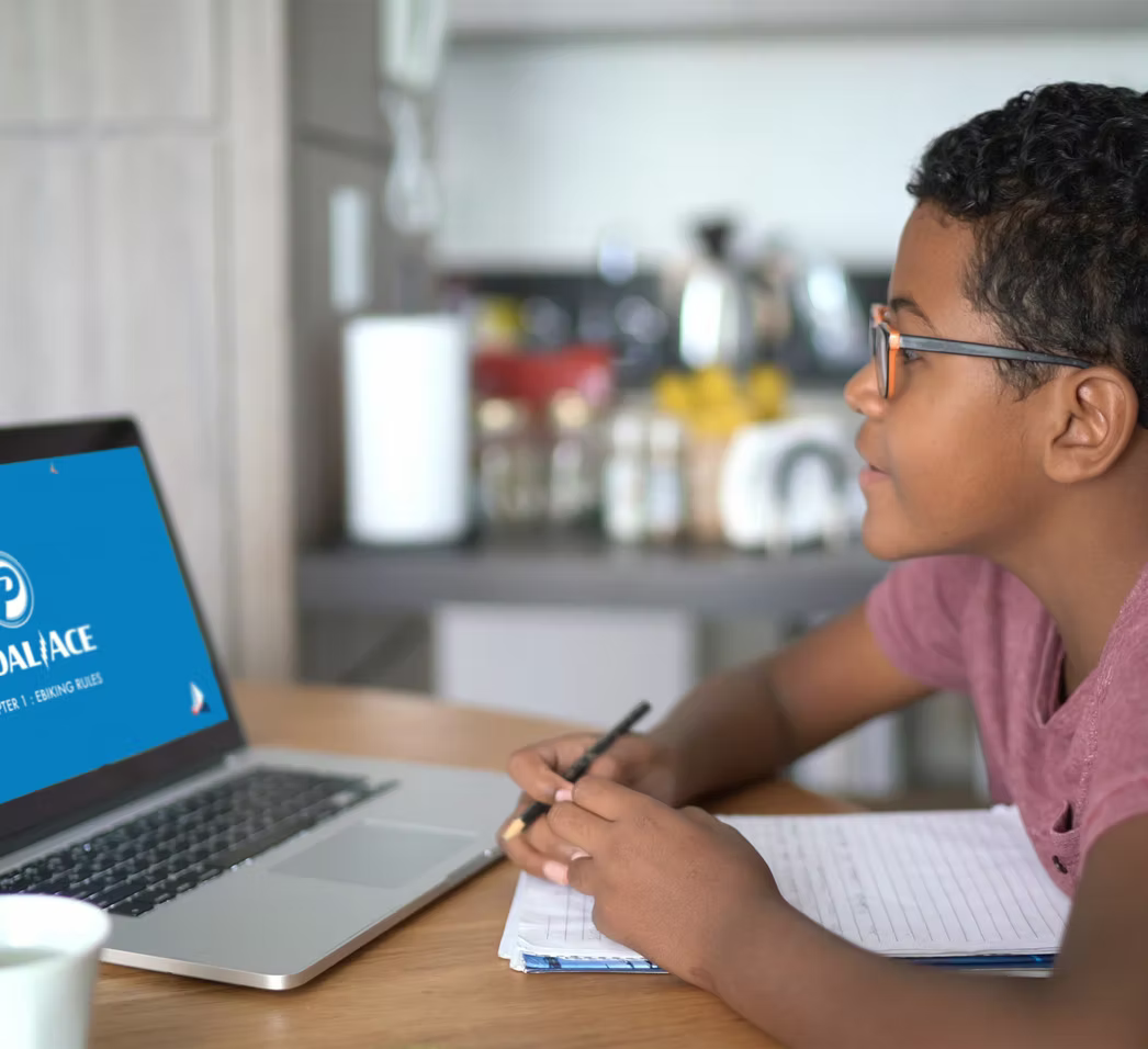 Child sitting at a table with a laptop and notebook, looking at the screen.