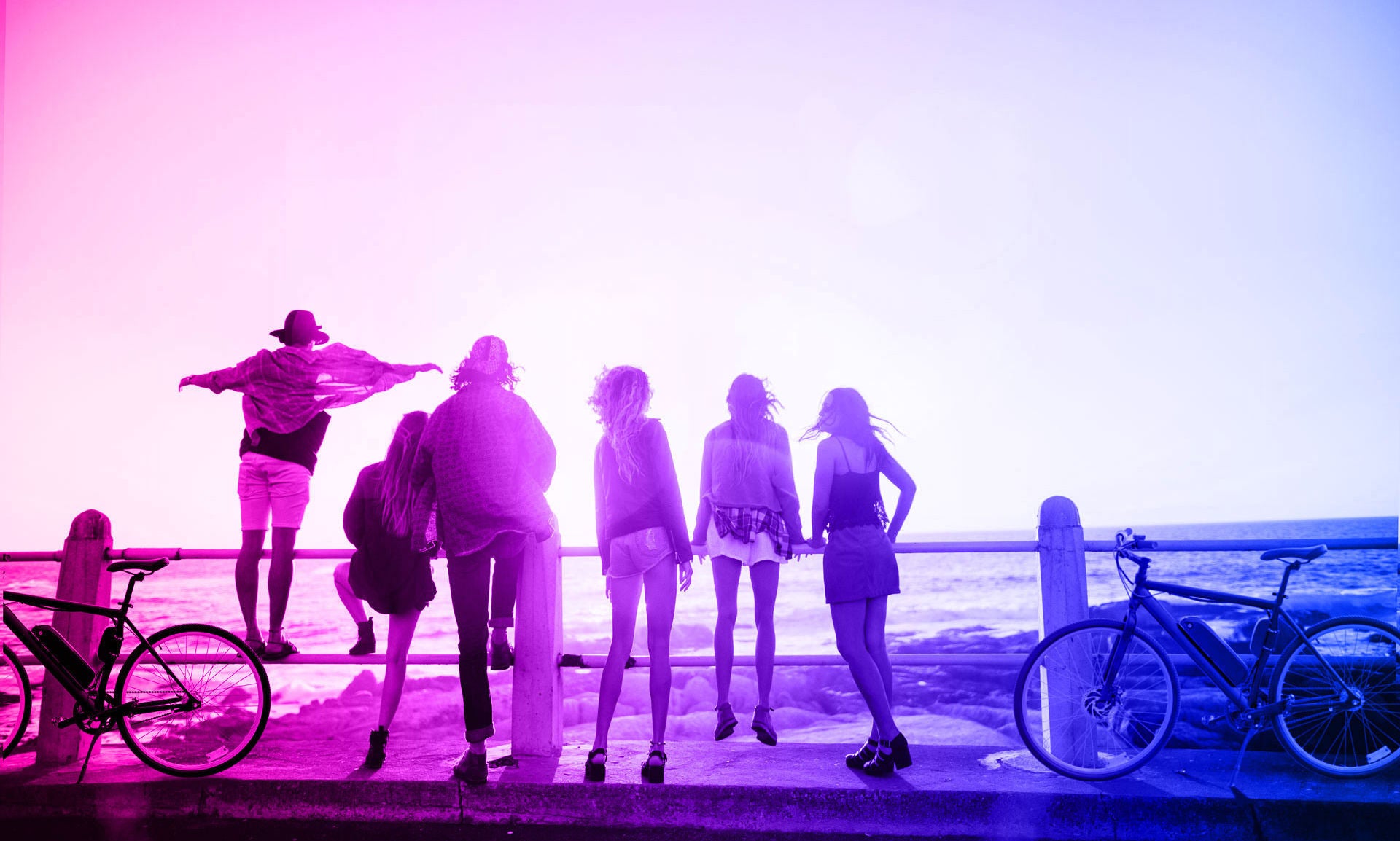 Group of people standing on a pier with bicycles, silhouetted against a colorful background.